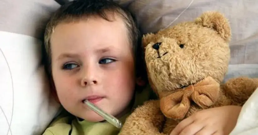 Chory chłopiec z termometrem w ustach i pluszowym misiem w łóżku.