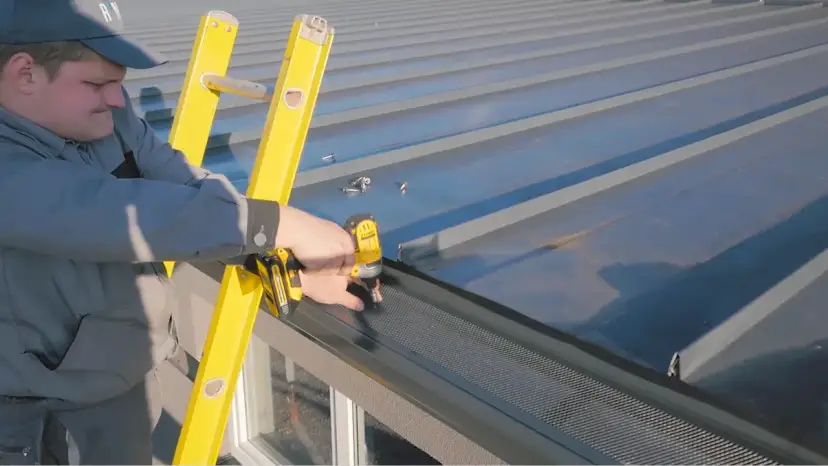 Dekarz przykręca siatkę na rynnie na dachu z blachy.