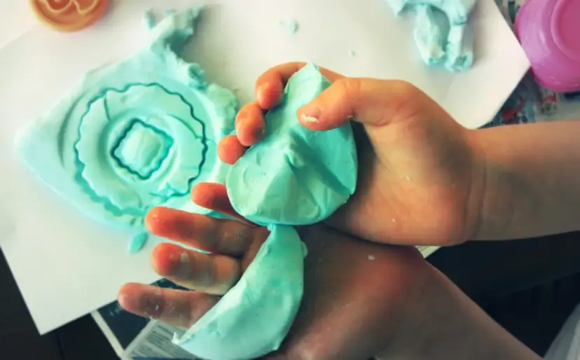 Dziecko bawi się niebieską ciastoliną, tworząc z niej różne kształty. Pokazane są ręce dziecka ugniatające masę, a obok leży uformowana z ciastoliny ozdobna forma. To świetna zabawa, jak zrobić taką ciastolinę?