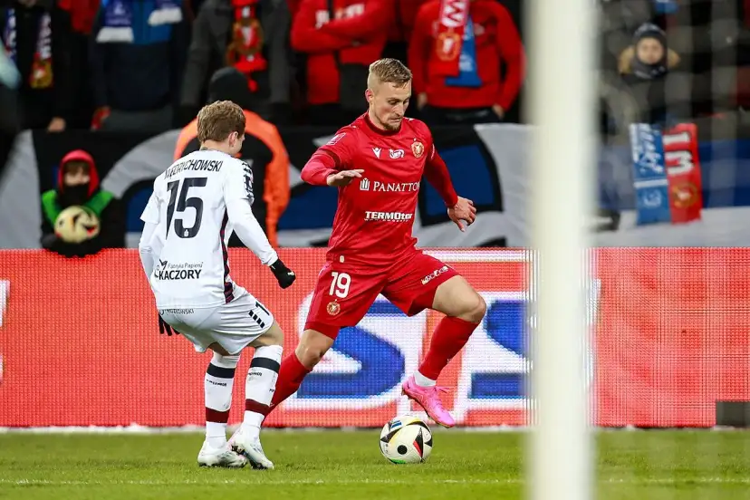 Piłkarze walczą o piłkę na boisku. Jeden w czerwonej koszulce z numerem 19, drugi w białej z numerem 15. To ważny mecz w sezonie Ekstraklasy.