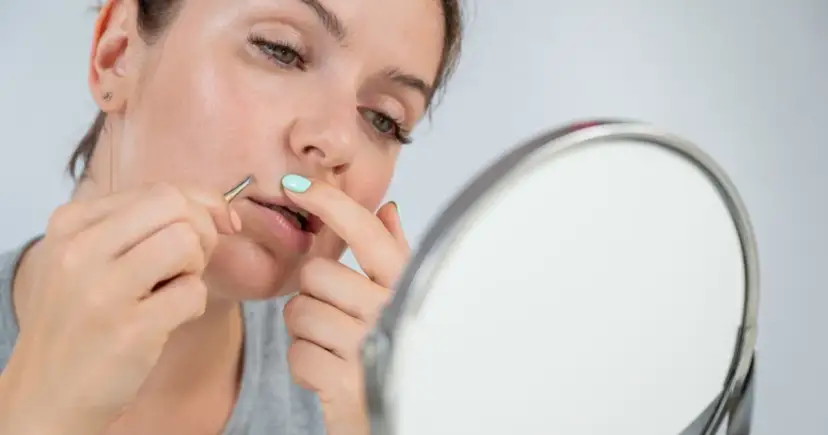 Kobieta wyrywa pęsetą włoski nad górną wargą, patrząc w lustro.