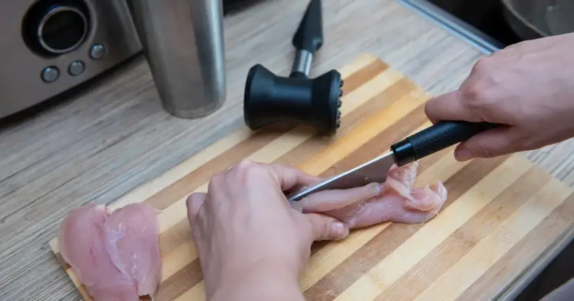 Ręce krojące surowego fileta z kurczaka na desce do krojenia.