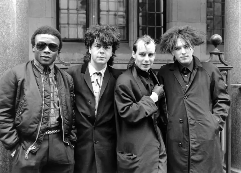 Vier Männer in dunkler Kleidung, Post-Punk-Ästhetik, stehen vor einem Gebäude.