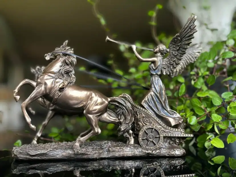Figurka Nike, bogini zwycięstwa, w rydwanie zaprzężonym w dwa konie, na tle zieleni.