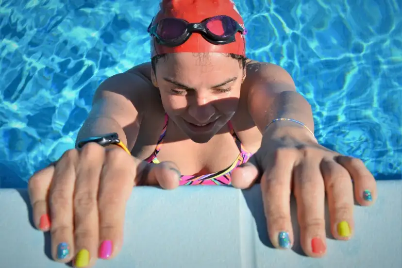 Kobieta w czerwonym czepku pływackim i goglach opiera się o brzeg basenu.