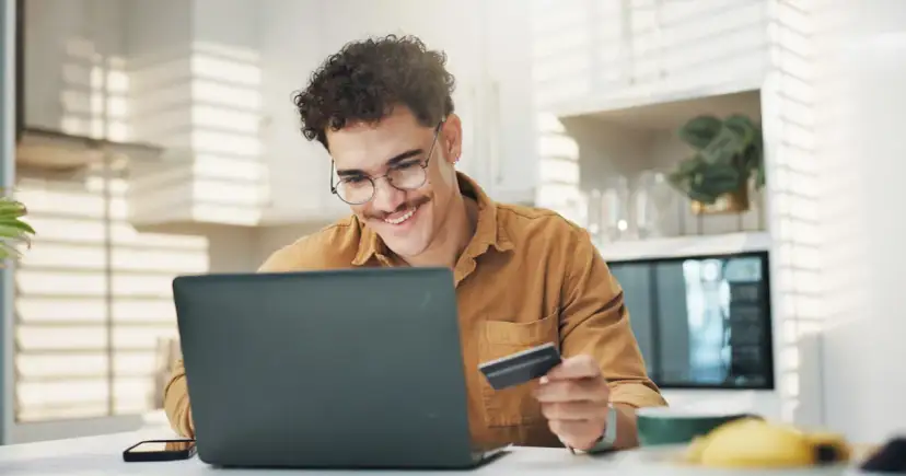 Uśmiechnięty mężczyzna z kartą płatniczą przed laptopem, być może sprawdza limit w koncie przed zakupami online.