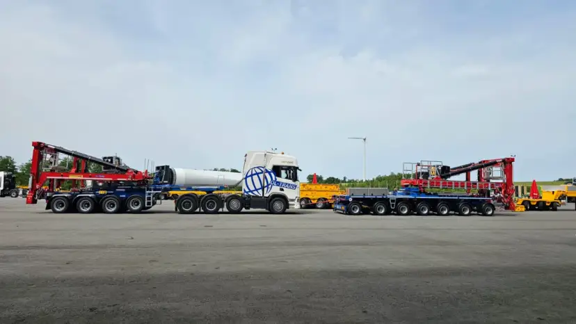 Ciężarówka z naczepą tandem przewozi element turbiny wiatrowej. Obok stoi inna naczepa z dźwigiem.