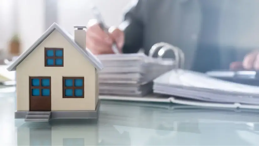 Model domu na biurku, obok stosy dokumentów i osoba pisząca. Czy kupując dom od dewelopera płacę podatek?