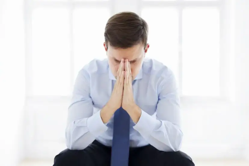 Mężczyzna w koszuli i krawacie z rękami złożonymi na twarzy, w zamyśleniu.