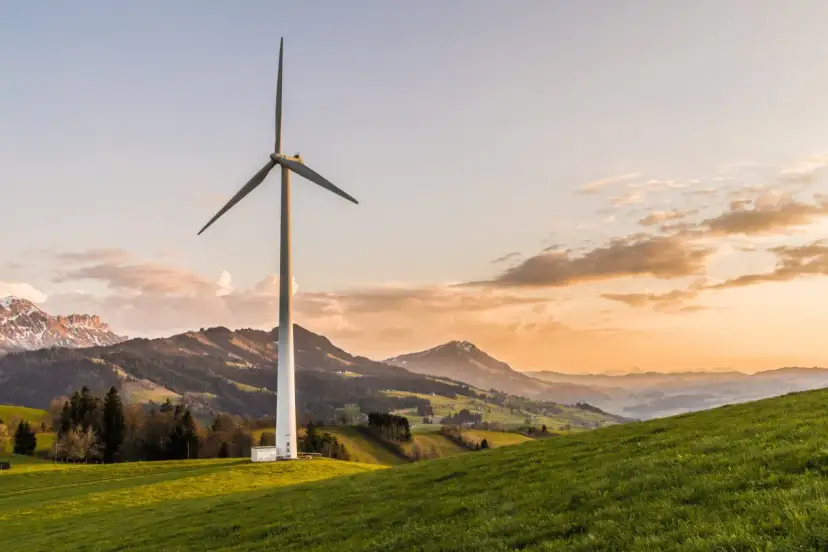 Wiatrak na zielonym wzgórzu, symbolizujący czystą energię z wiatru, na tle malowniczych gór o zachodzie słońca.