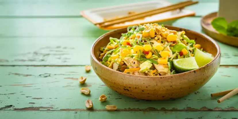 Kolorowa sałatka tajska z kurczakiem, kukurydzą, orzeszkami ziemnymi i limonką w glinianej misce na zielonym, drewnianym stole.