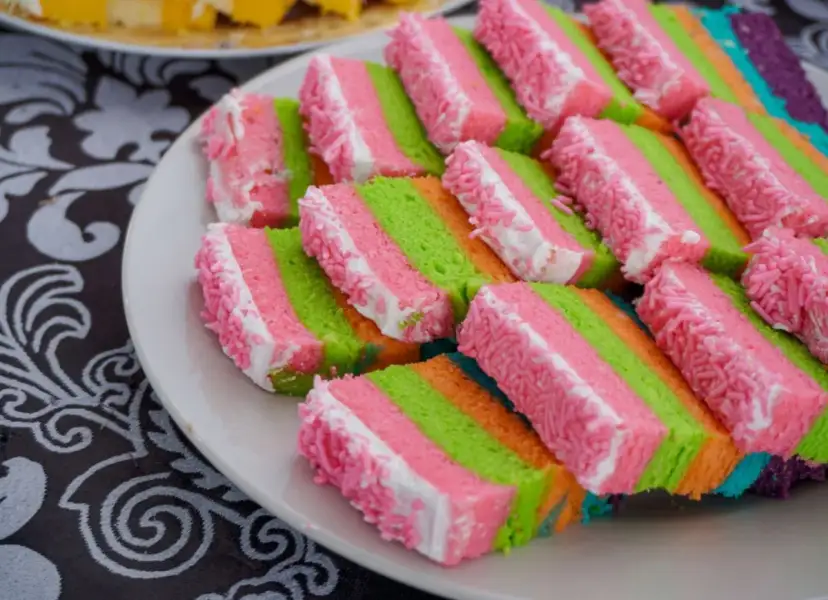 Kolorowe ciasto dla dzieci do przedszkola, pokrojone w paski różowe, zielone i pomarańczowe, ozdobione białym kremem i różową posypką.