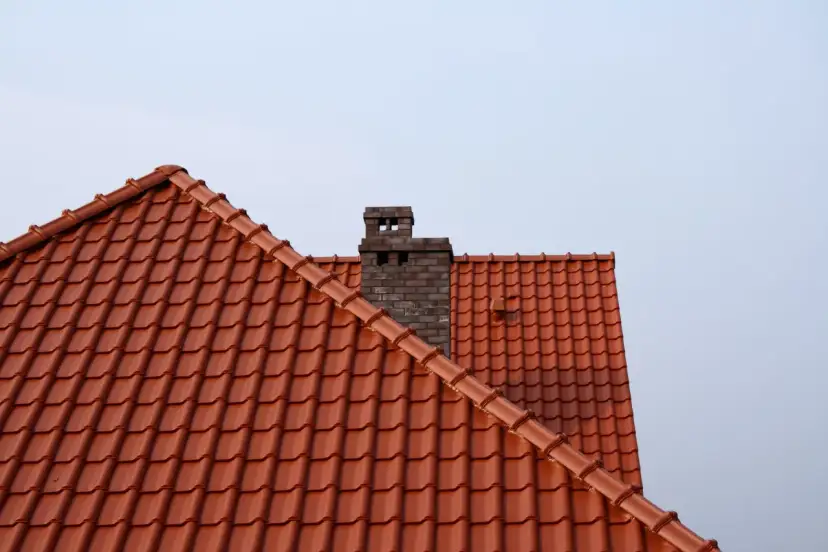 Ceglany komin na tle jasnego nieba, widok na dach pokryty czerwonymi dachówkami.