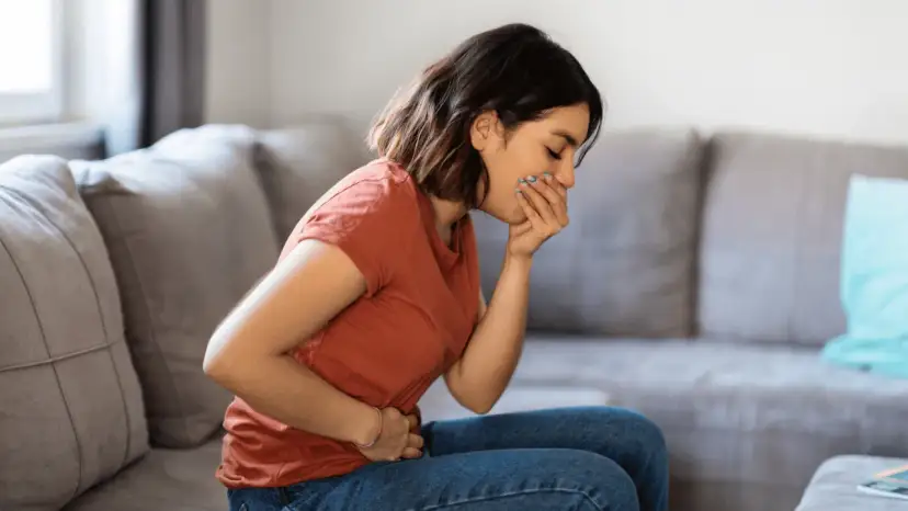 Kobieta siedzi na kanapie, trzymając się za brzuch i zakrywając usta, odczuwając mdłości.