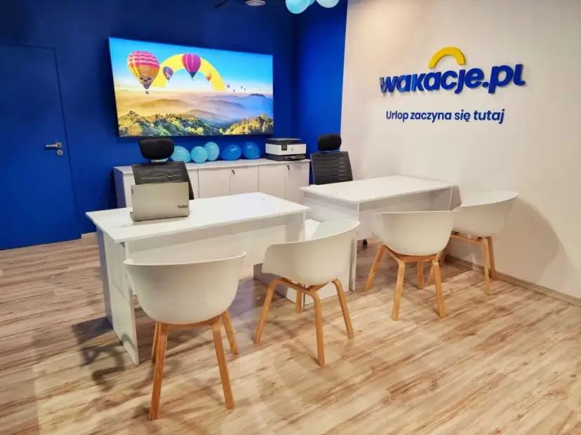Nowoczesne biuro z logo "wakacje.pl", ekranem z balonami i białymi biurkami.