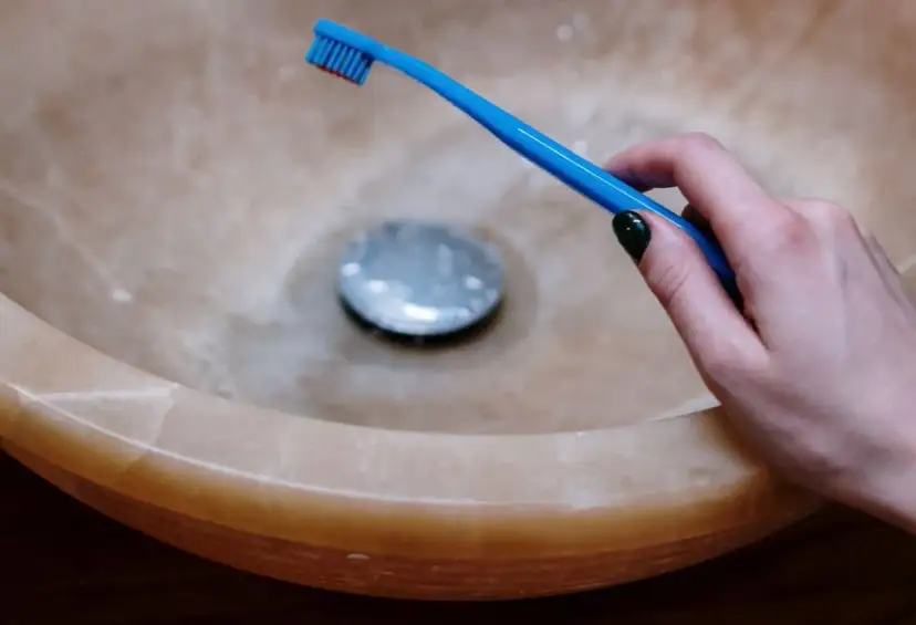 Dłoń trzymająca niebieską szczoteczkę do zębów nad umywalką. Pytanie brzmi: jak długo myć zęby?