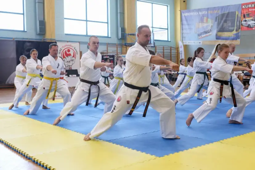 Grupa osób w kimonach ćwiczy karate na niebiesko-żółtej macie w sali gimnastycznej.
