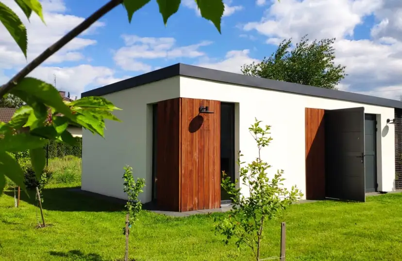 Nowoczesny biały budynek gospodarczy z drewnianymi elementami i otwartymi drzwiami na zielonym trawniku.