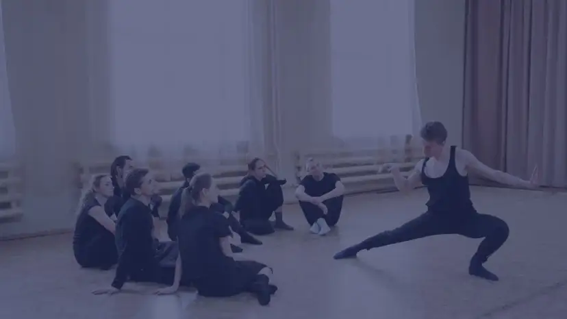 Choreograf demonstruje ruchy grupie tancerzy siedzących na podłodze.