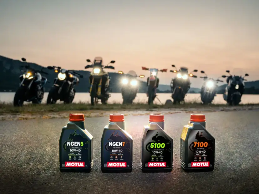 Cztery butelki oleju Motul do motocykla: NGEN5, NGEN7, 5100 i 7100, stoją na drodze o zachodzie słońca, za nimi widać rzędem motocykle.