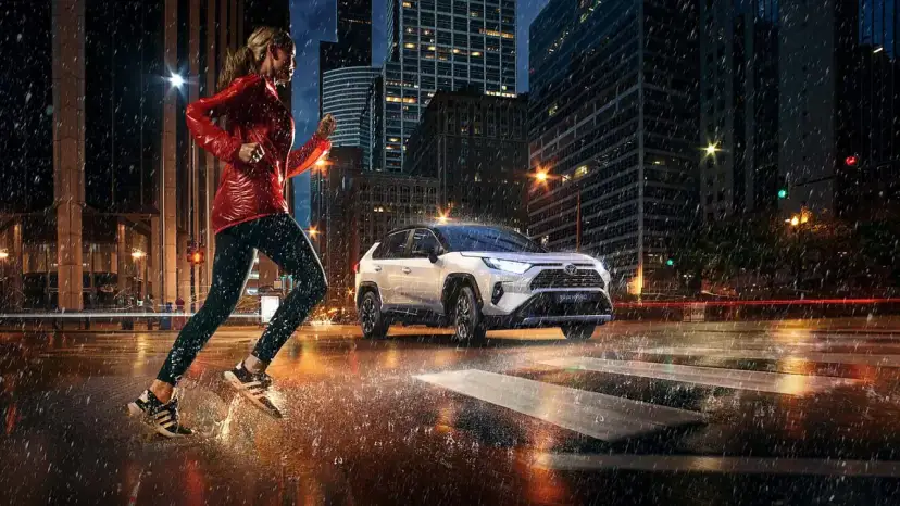Biegaczka w deszczowy wieczór w mieście, obok niej biała Toyota RAV4 Hybrid. Zastanawiasz się, ile pali Toyota RAV4 Hybrid?