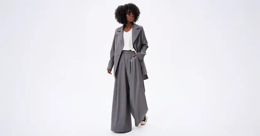 Kobieta w szarym garniturze z szerokimi spodniami i marynarką na białym tle.