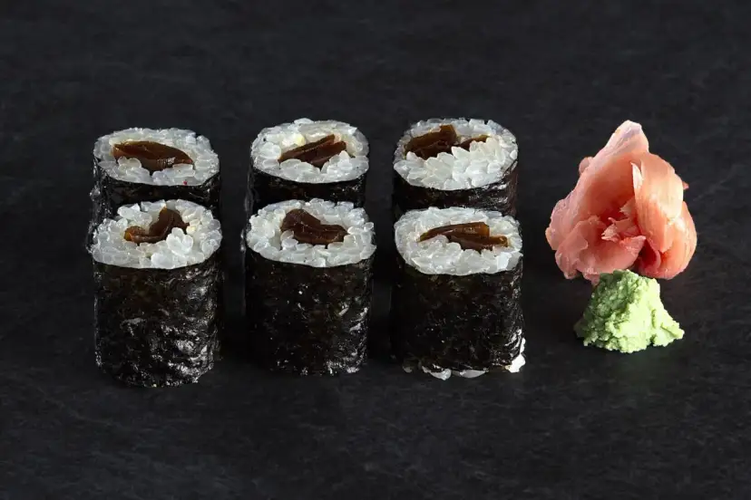 Sześć roladek sushi z ryżem i ciemnym nadzieniem, prawdopodobnie sushi z tykwą. Obok różowy imbir i zielone wasabi.