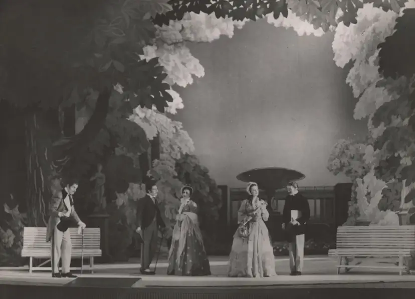 Scena z przedstawienia teatralnego, gdzie aktorzy w strojach z epoki odgrywają scenę w ogrodzie. Reżyser teatralny Szyfman zadbał o szczegóły scenografii.