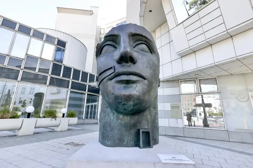 Brązowa rzeźba głowy Igora Mitoraja na tle nowoczesnego budynku.