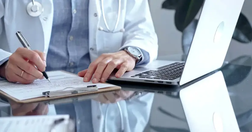 Lekarz w białym kitlu pisze na dokumentach, obok laptop.