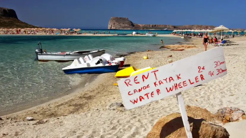 Maj na Krecie: plaża z leżakami, turkusowa woda, łódki i tablica z ofertą wynajmu kajaków i rowerów wodnych.