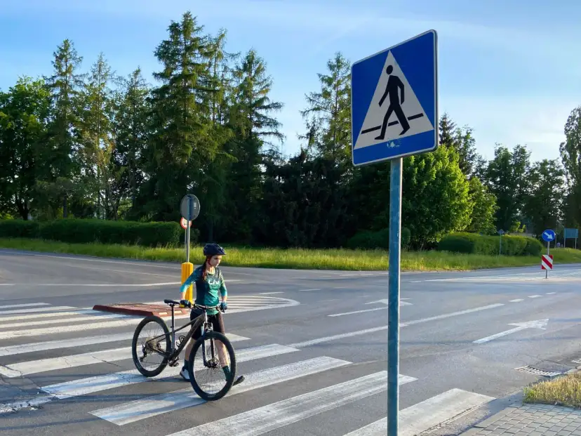 Rowerzystka w kasku prowadzi rower po pasach dla pieszych obok znaku drogowego.