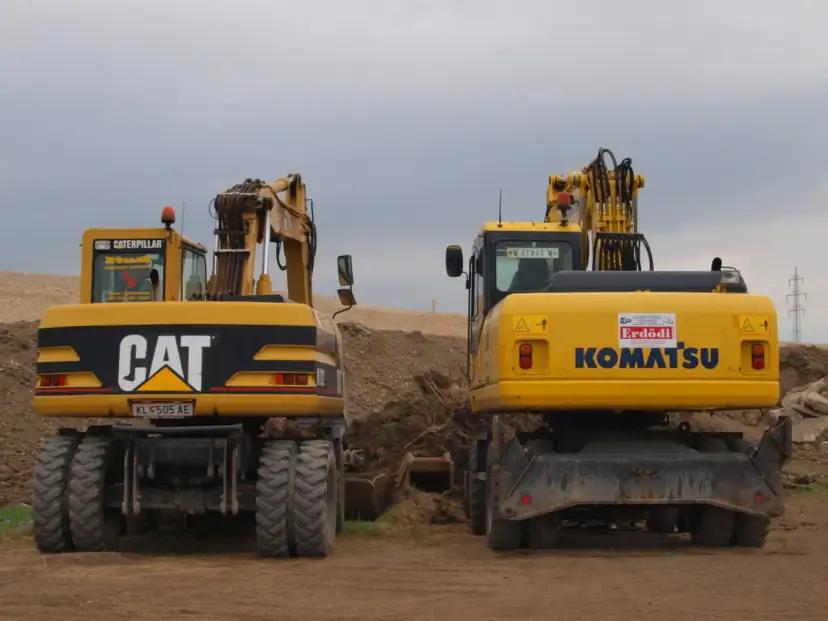Dwie żółte koparki, CAT i Komatsu, stoją na placu budowy. Czy koparka ma dowód rejestracyjny? Na tablicy rejestracyjnej CAT widnieje KL 505 AE.