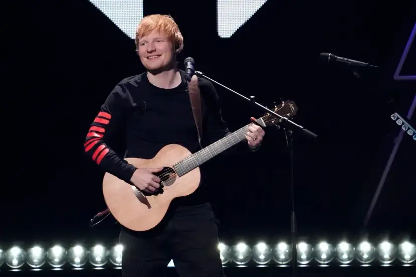 Ed Sheeran gra na gitarze akustycznej na scenie, ubrany w czarną bluzę z czerwonymi pasami na rękawach.