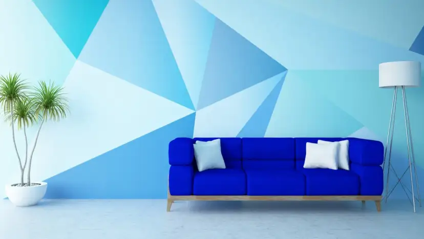 Niebieska sofa stoi przed ścianą pomalowaną do połowy w geometryczne wzory. Obok stoi lampa i roślina.