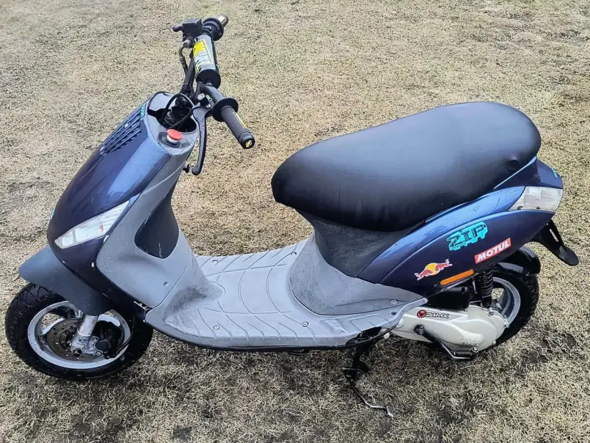 Niebieski skuter z naklejkami Red Bull i Motul na tle trawy.