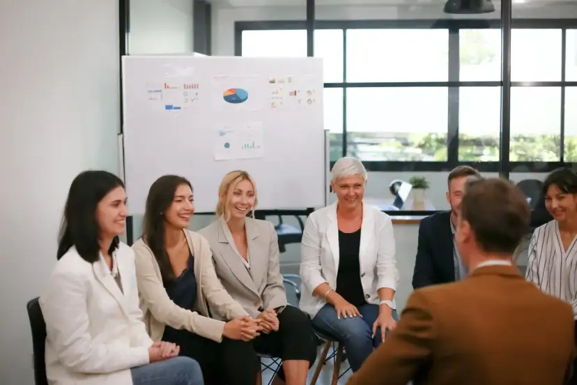 Grupa ludzi siedzi w kręgu, uśmiechając się i rozmawiając. Na ścianie w tle wiszą wykresy.