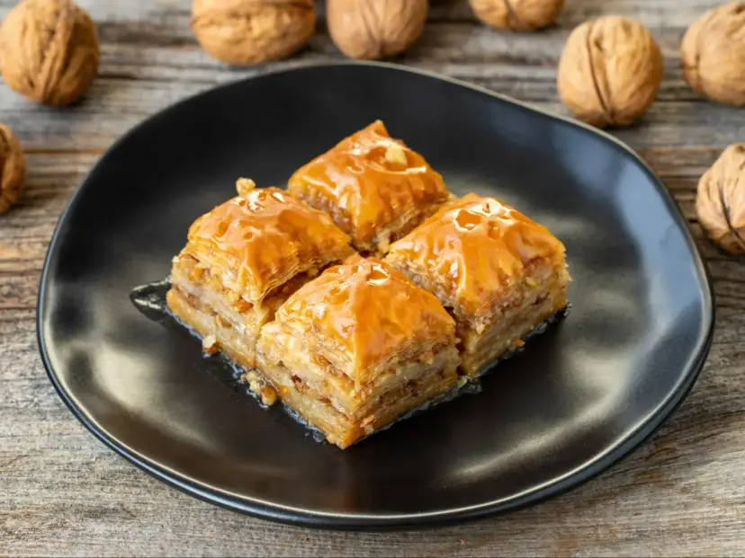 Cztery kawałki baklawy na czarnym talerzu, polane syropem. W tle orzechy włoskie. Idealny przepis na baklavę.