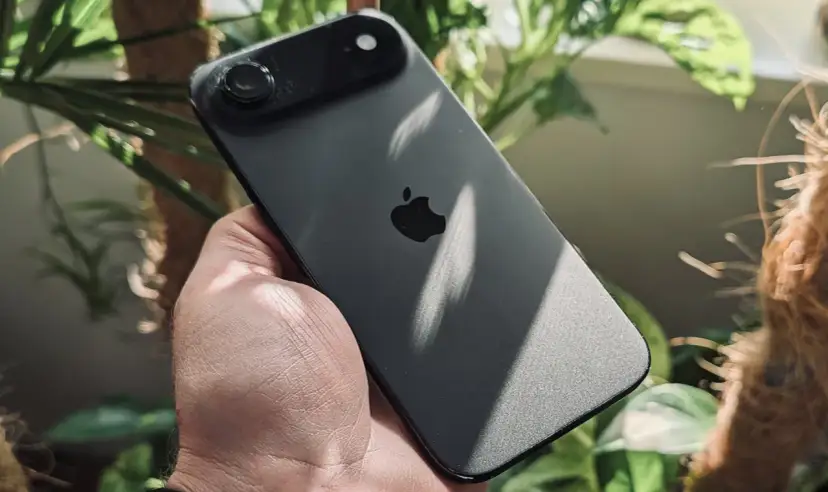 Ciemny iPhone z jednym aparatem w dłoni, otoczony zielenią roślin.