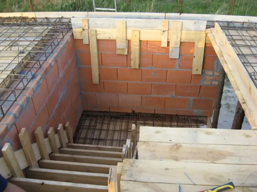 Budowa schodów dywanowych z betonowym zbrojeniem. Widać ceglane ściany, drewniane szalunki i siatkę zbrojeniową.
