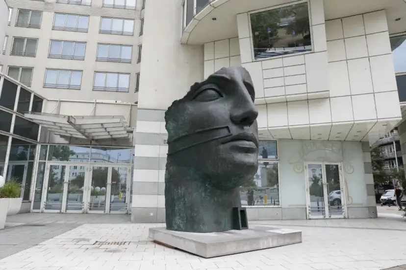 Okaleczona, ciemna rzeźba głowy z opaską na ustach, na tle nowoczesnego budynku.