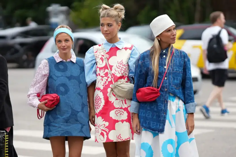 Trzy dziewczyny prezentują modny styl ubioru młodzieży: sukienki w kwiaty, dżinsowe kamizelki i kolorowe torebki.