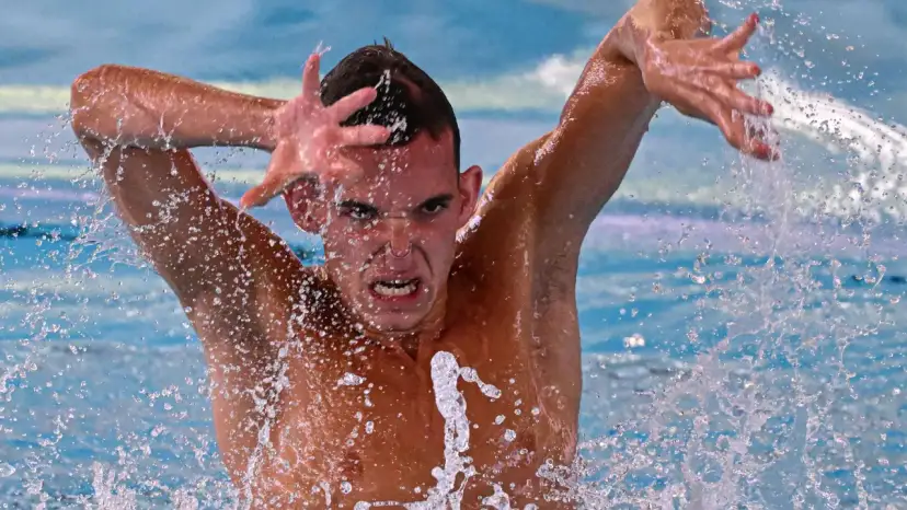 Mężczyzna wykonuje dynamiczny ruch podczas pływania synchronicznego, z twarzą skupioną i wodą rozpryskującą się wokół.
