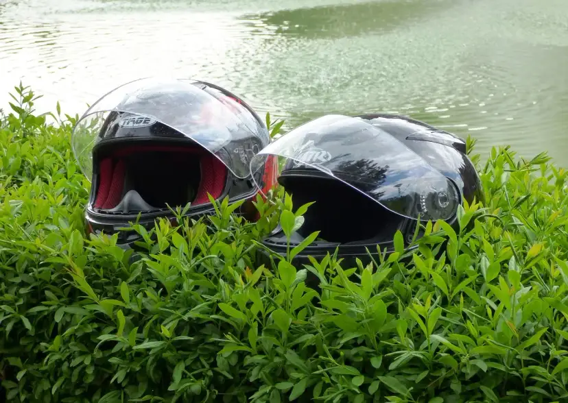 Dwa kaski motocyklowe w zielonych liściach, z wodą w tle.