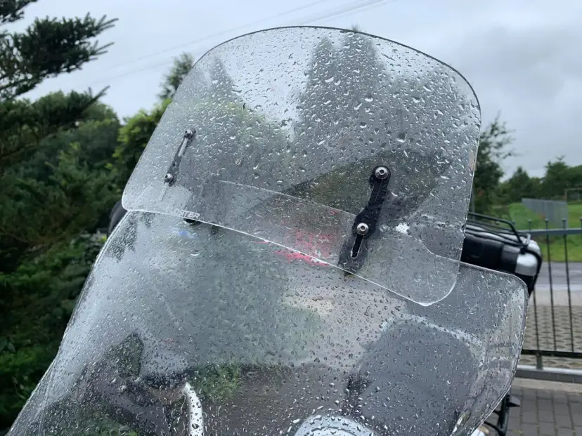 Przednia szyba motocykla pokryta kroplami deszczu, z widocznymi mocowaniami.