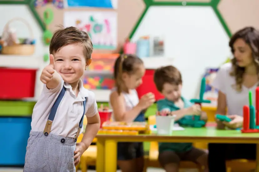Uśmiechnięty chłopiec pokazuje kciuk w górę w przedszkolu, gdzie od kiedy zerówka jest obowiązkowa, dzieci bawią się i uczą.