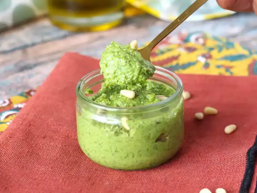 Łyżka nabiera aromatyczne pesto z rukoli z małego słoiczka. Na czerwonym tle widać orzeszki piniowe.
