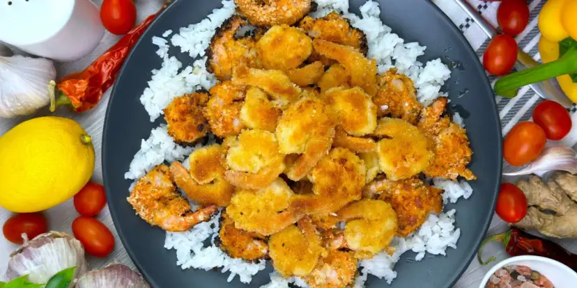 Chrupiące krewetki w złocistej panierce do krewetek, podane na białym ryżu. Obok cytryna, pomidorki i przyprawy.