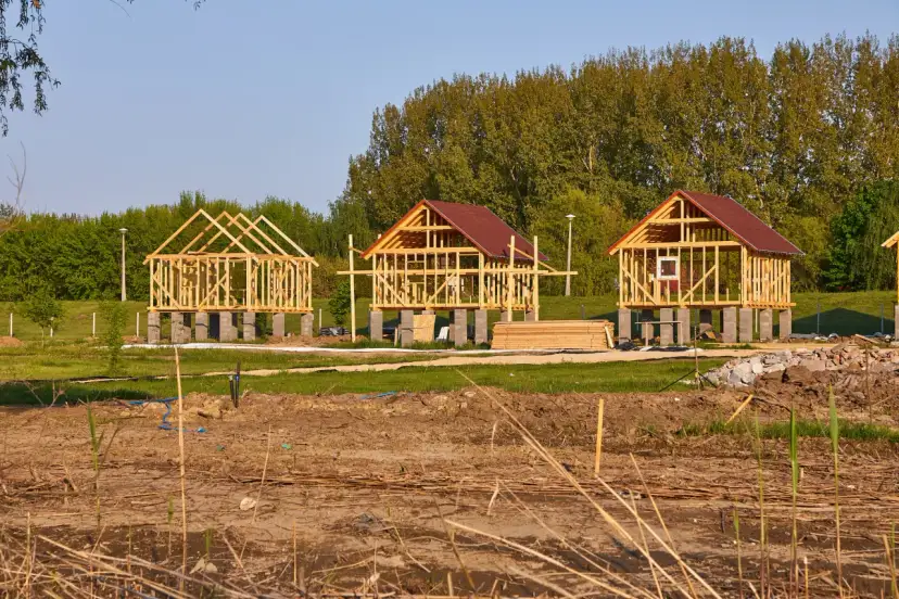 Trzy drewniane szkielety budynków gospodarczych na betonowych fundamentach, w tle drzewa.