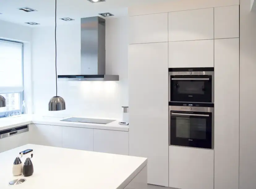 Nowoczesna kuchnia z białymi frontami do szafek kuchennych, stalowym okapem i wbudowanymi piekarnikami.